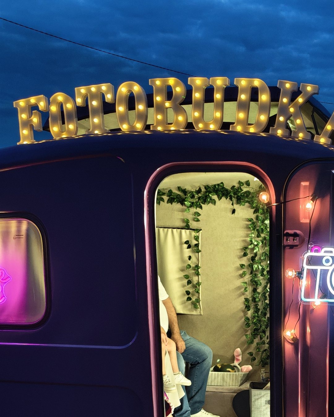 Fotofurka wieczorem – podświetlony szyld FOTOBUDKA, neon aparatu i otwarte drzwi przyczepy