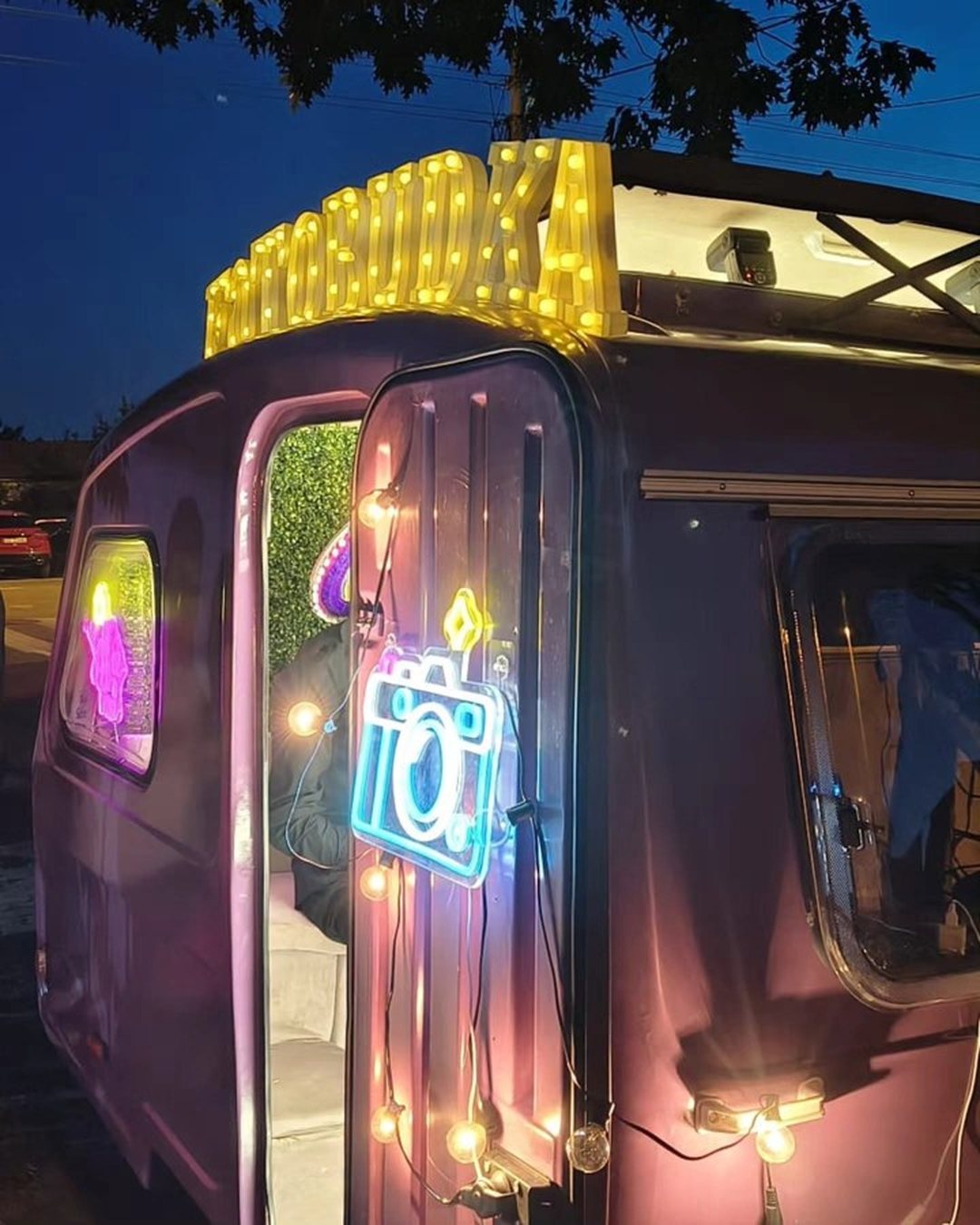 Neonowy szyld FOTOBUDKA i neon aparatu na fioletowej przyczepie nocą
